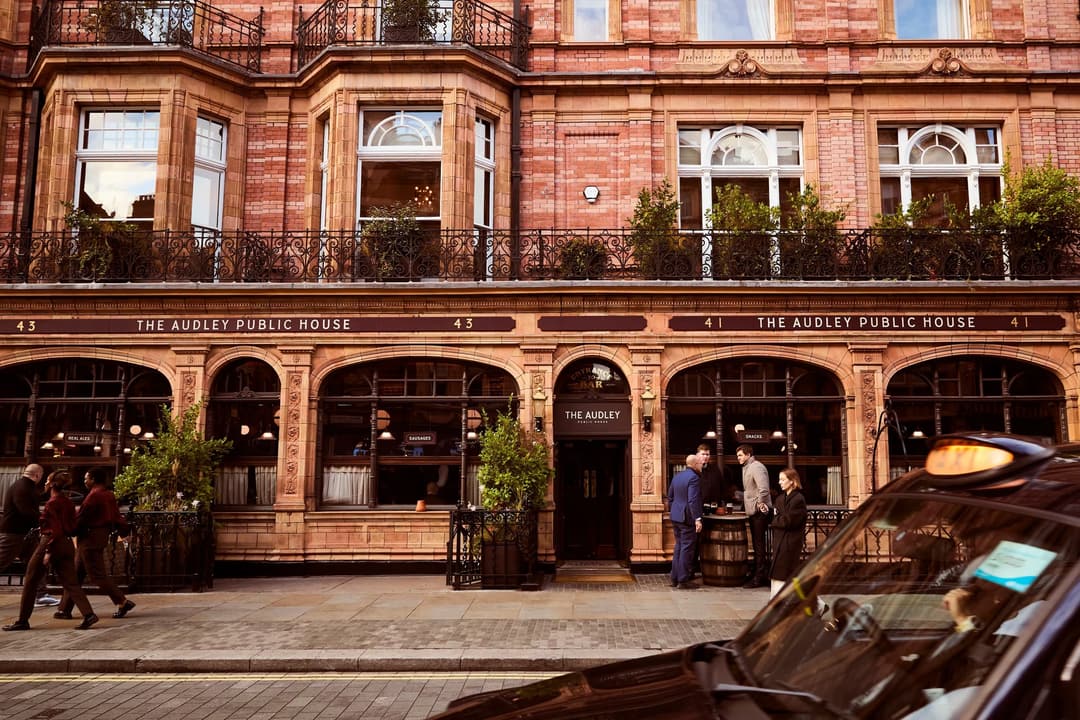 The Audley Public House in a red-brick Victorian building, with pedestrians outside and a black taxi passing in the foreground, The Connaught