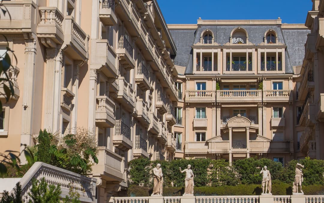 Elegant multi-story beige building with ornate balconies and classical statues in front under clear blue sky.