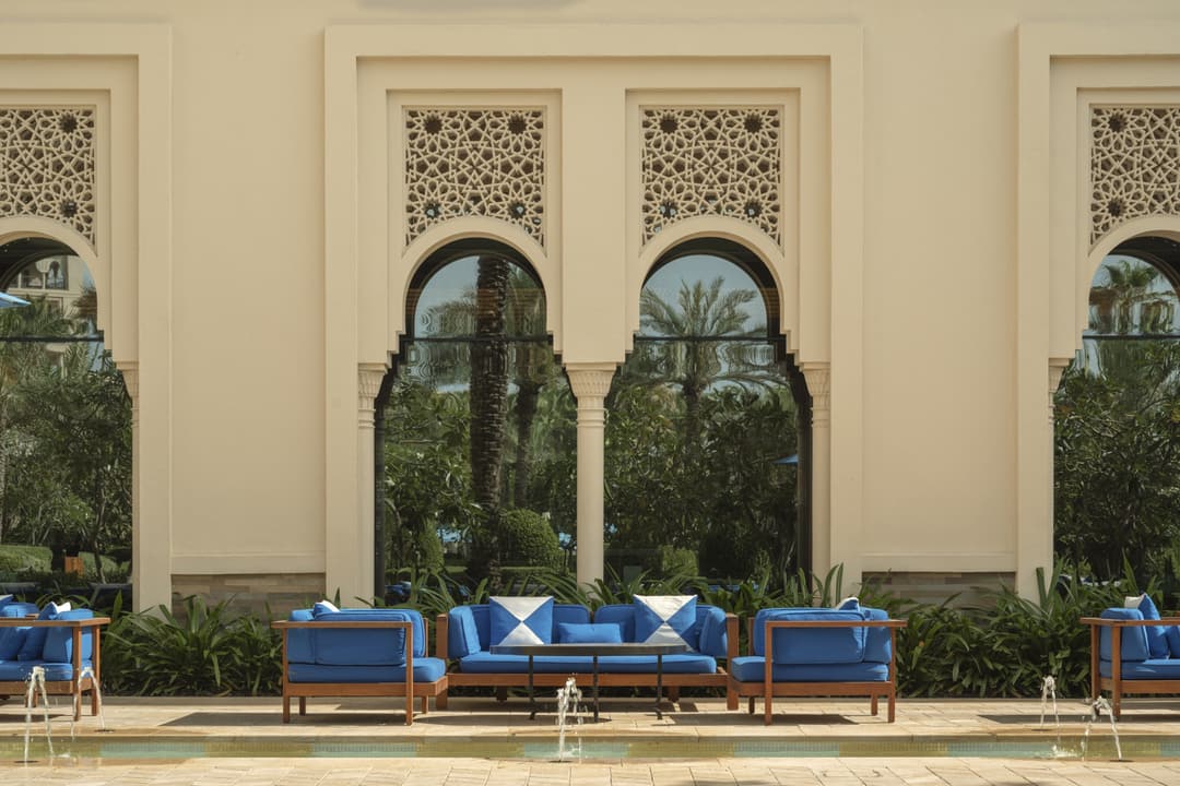 A sunny courtyard with upholstered seating and greenery, with an exterior hotel wall with tall, elegant arched windows in the background, Four Seasons