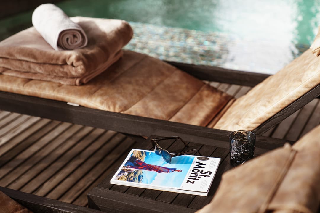 Poolside lounge chair with beige cushions, rolled towels, magazine, sunglasses, and water glass beside turquoise pool, Carlton Hotel St. Moritz