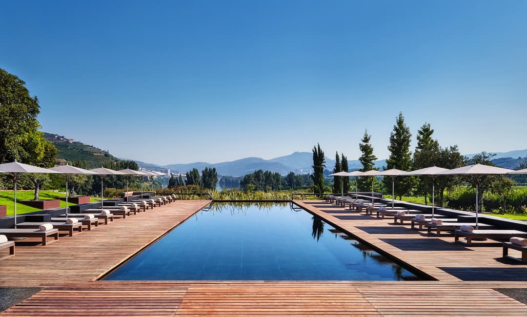 A tranquil pool area with lounge chairs and umbrellas on wooden decks, surrounded by lush vegetation and distant mountains under a blue sky, Six Sense