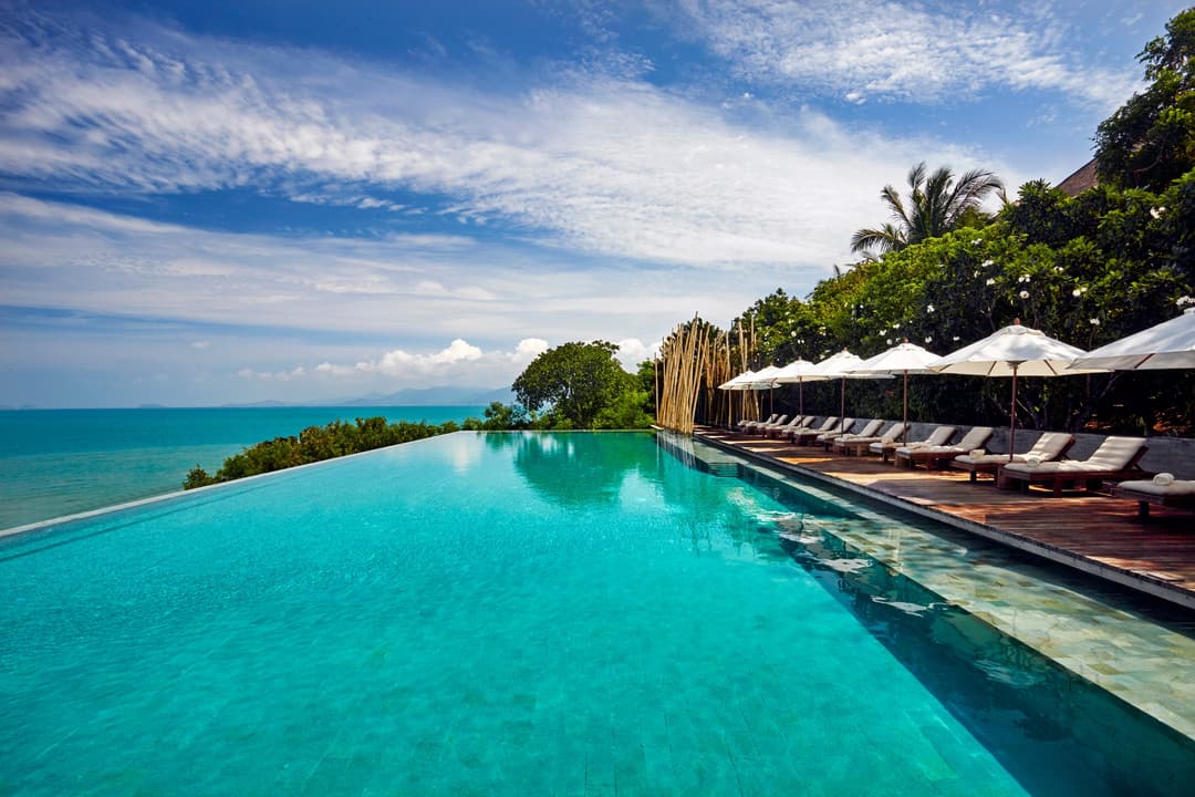 Oceanview pool with accessible loungers and shaded areas on a wooden deck, bordered by native greenery beneath clear skies, Six Senses Samui