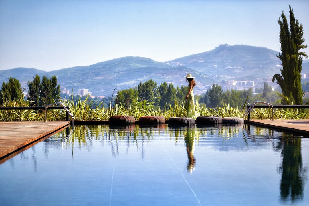 A guest strolls beside a tranquil swimming pool bordered by lush gardens, with gentle hills and a distant city skyline beneath an open sky, Six Senses