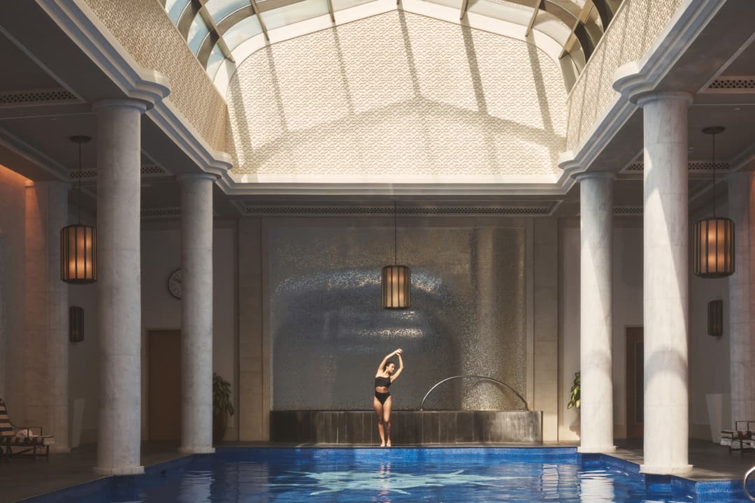 Person in swimsuit standing and stretching with arms overhead beside indoor pool flanked with white pillars under vaulted ceiling, Four Seasons Resort