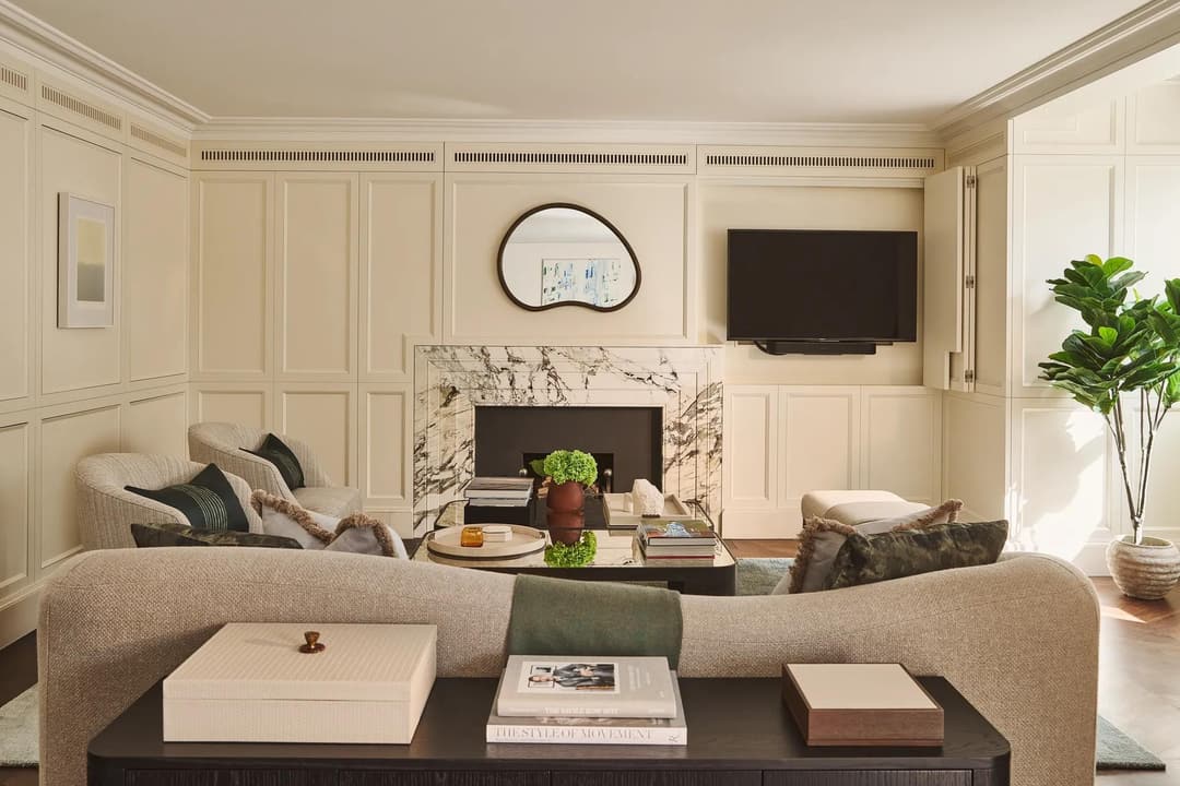 Bright living room with marble fireplace, curved sofa, armchairs, wall-mounted TV, and coffee table, The Connaught