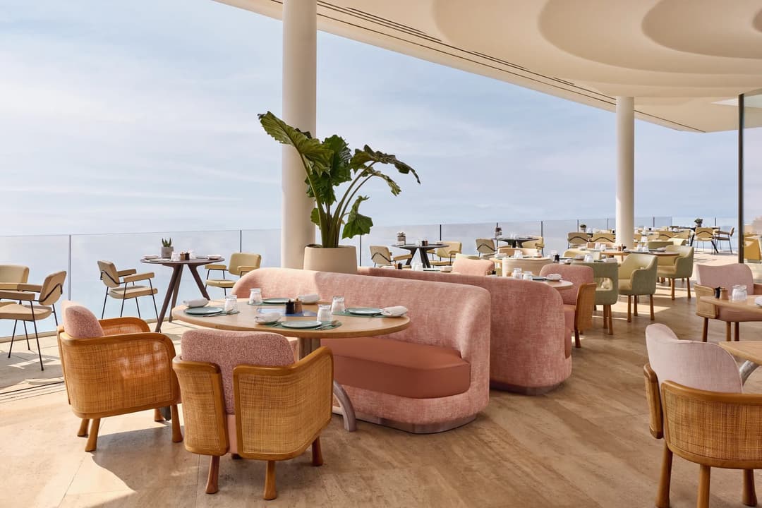 Elegant restaurant interior with pink sofas, wooden chairs, and sea views through floor-to-ceiling windows, featuring soft natural light and modern décor.