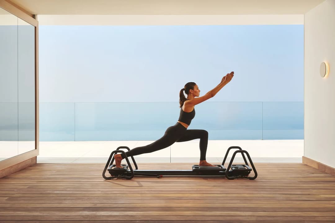 Woman practicing Pilates on a reformer in a modern studio with ocean view through glass walls.