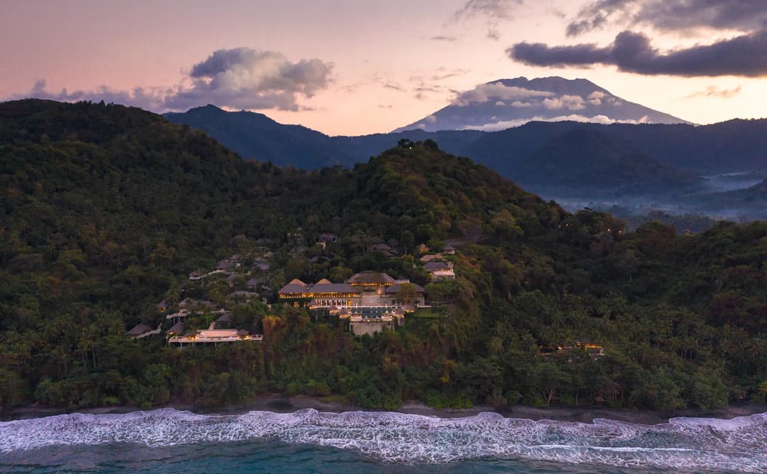 Aerial view of Amankila at dusk, with forested hills descending to a crescent beach and calm waters beyond