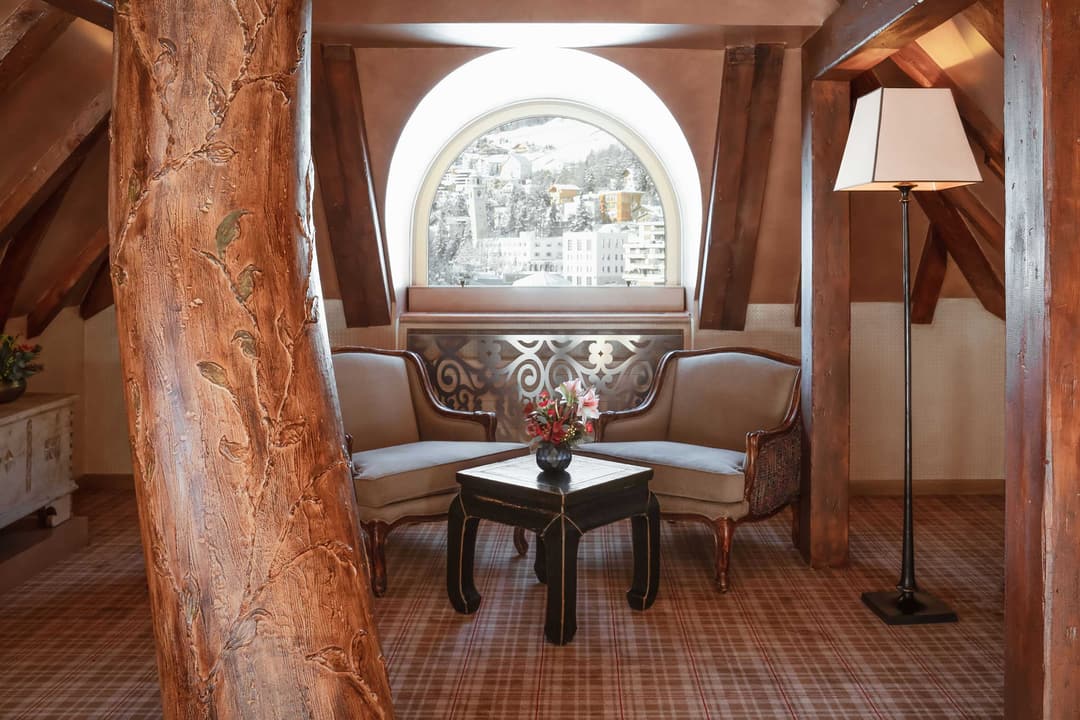 Cozy attic sitting area with exposed wooden beams, two beige armchairs, arched window overlooking snowy mountain village, Carlton Hotel St. Moritz
