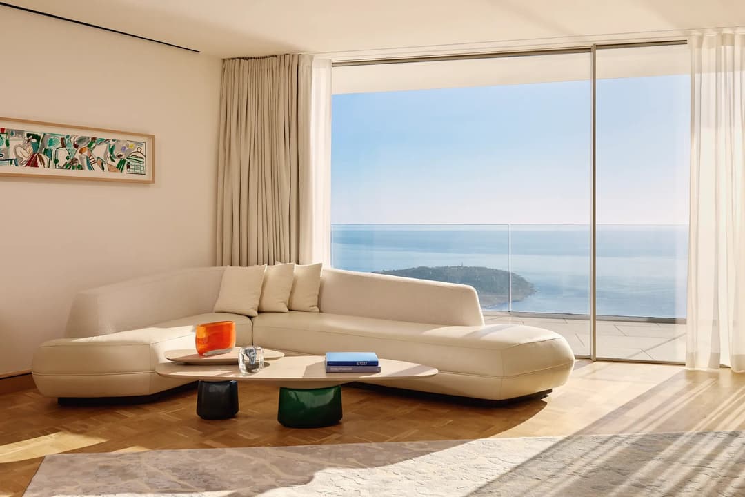 Canapé crème aux courbes sculpturales face à des baies vitrées. L'océan et une péninsule boisée s'étendent à l'horizon. Parquet en chêne, œuvre abstraite, rideaux légers encadrent la vue.