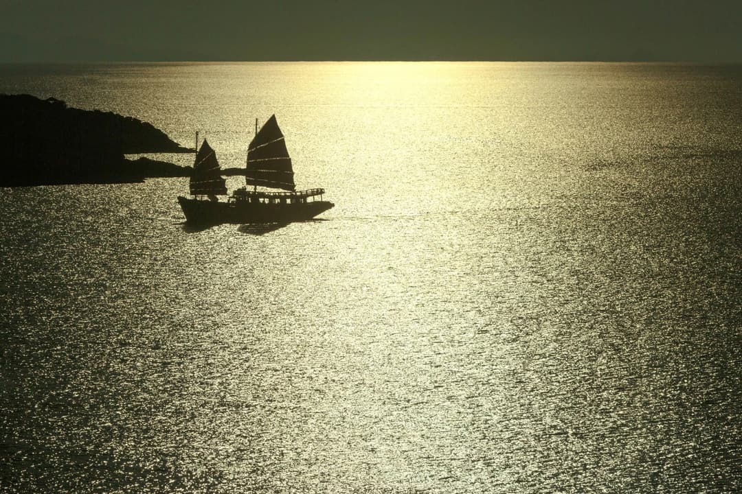 A sailboat with three sails moves across golden water at sunset, close to a calm coastline, reflecting a peaceful natural setting, Six Senses Samui