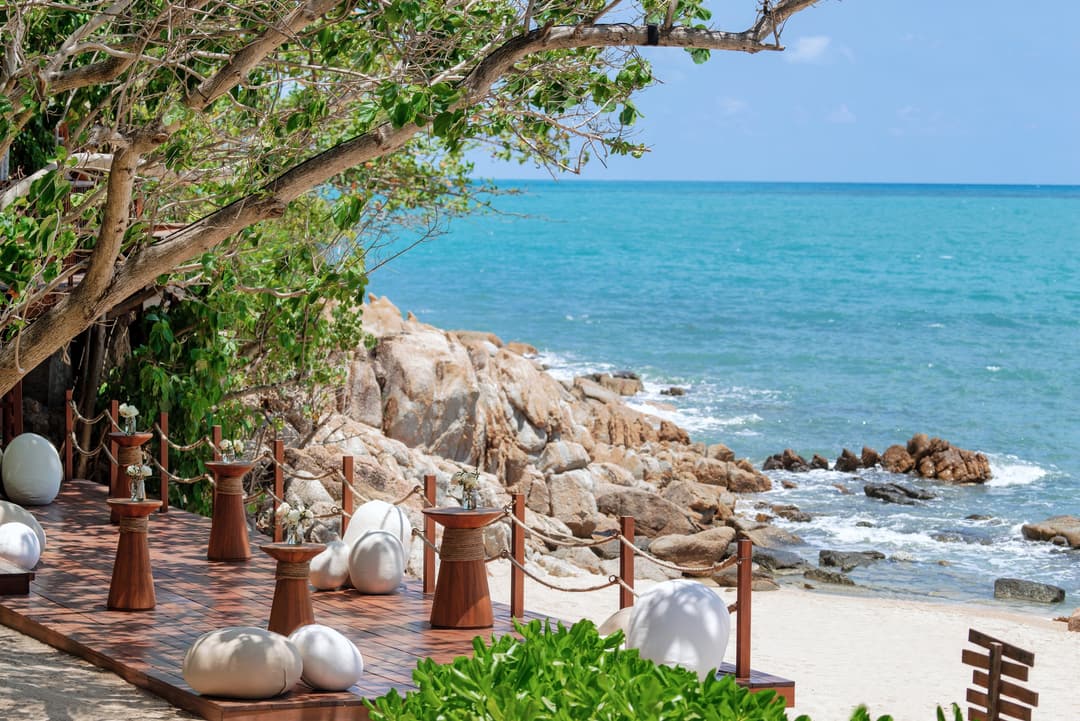 Wooden stools and white beanbags arranged on a deck by the beach, facing turquoise water and a rocky shore with an open sky above, Six Senses Samui