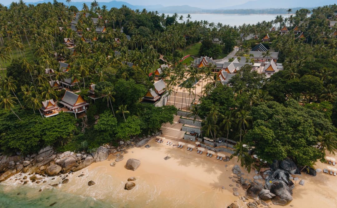 Aerial view of a sandy beach cove at Amanpuri, Thailand, surrounded by dense tropical forest and rocky outcrops