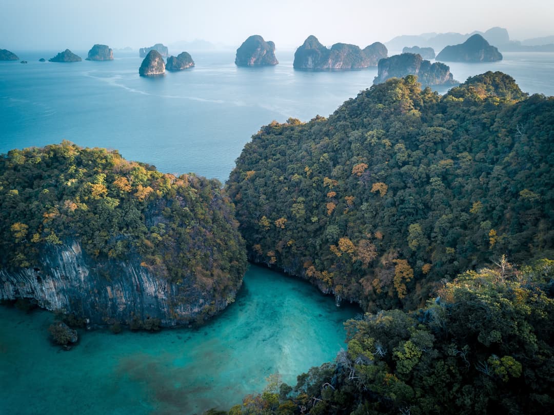 Forested islands framed by turquoise water, with rocky islets in the distance and a gentle, misty sky overhead, Six Senses Yao Noi