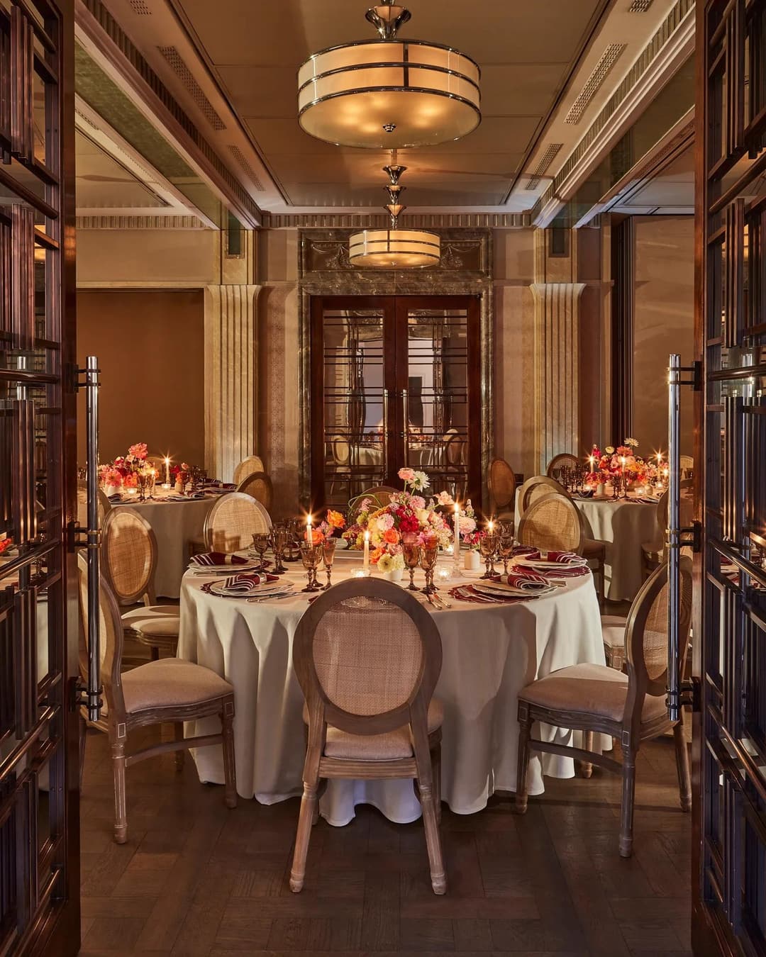 Warmly lit dining room with round tables set with floral centerpieces and candles, surrounded by elegant wooden chairs, The Connaught