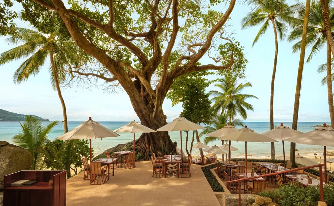 Dining terrace at Amanpuri with wooden tables set beneath palm trees overlooking the Andaman Sea