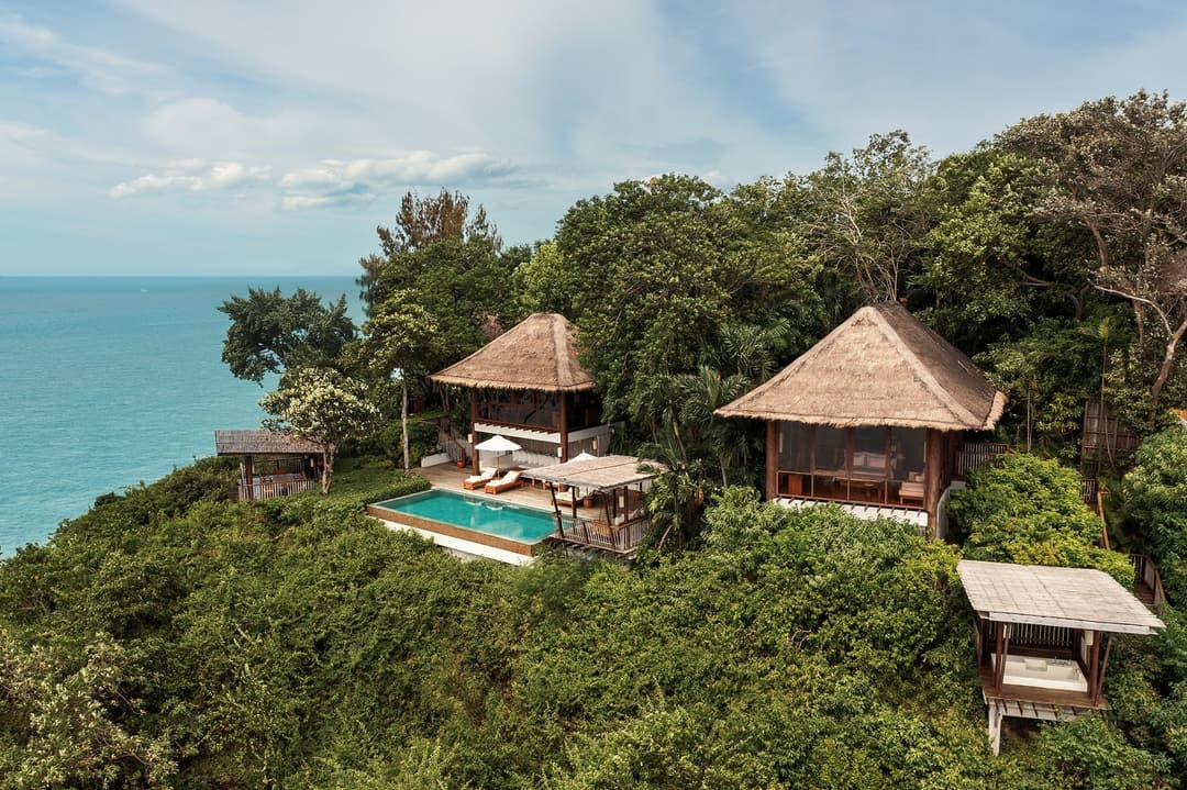 Villa with thatched roofs, private pool, and open lounge spaces set amid greenery, facing the blue ocean under a sky with scattered clouds, Six Senses