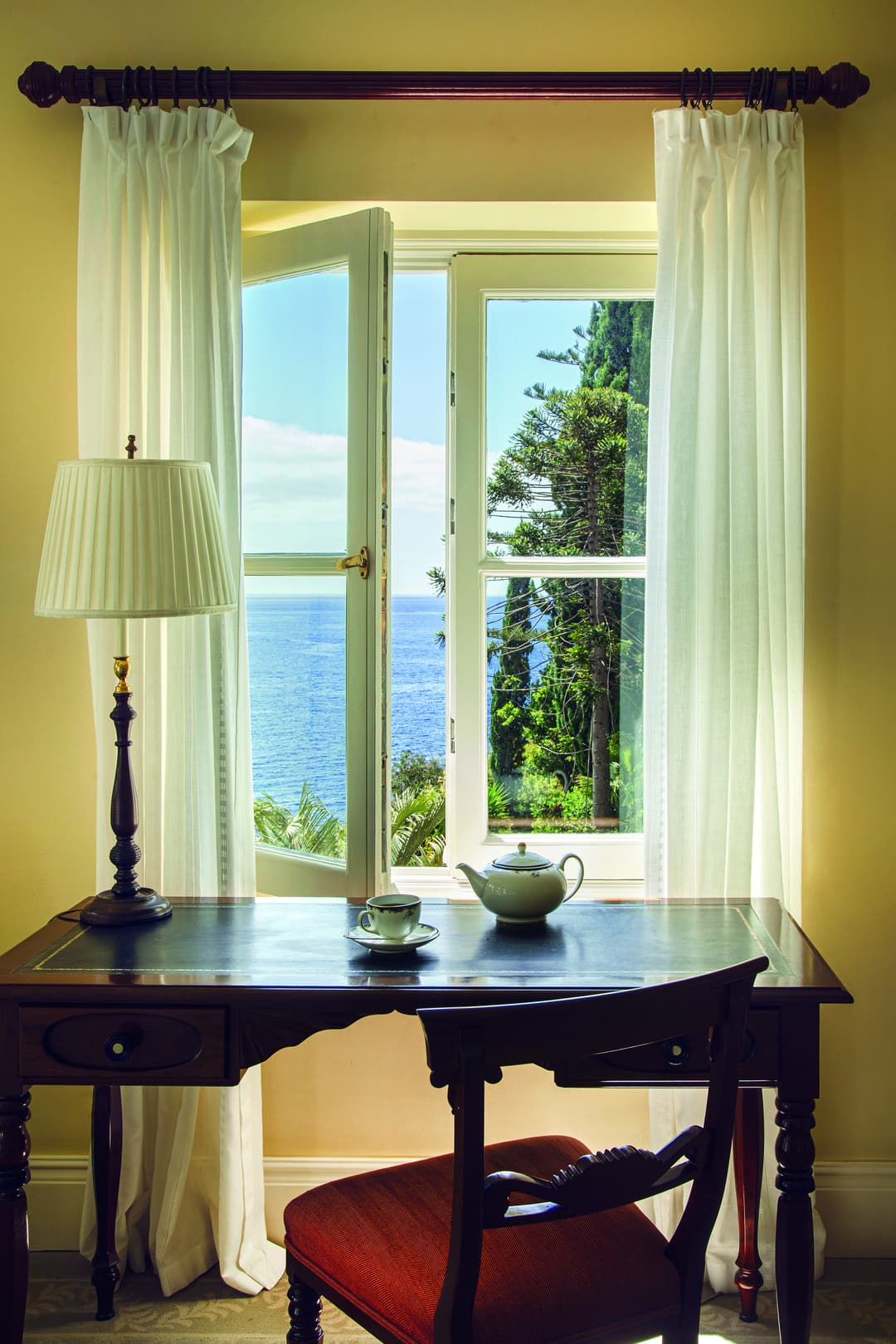 An elegant antique wooden desk with a lamp and a pot of tea by an open window with views of the botanical gardens and sea, Reid's Palace, A Belmond Ho