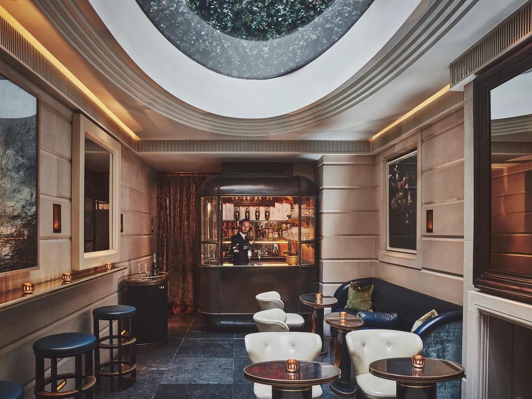 Intimate bar lounge with round tables, leather seating, and a skylight above, facing a small illuminated bar counter, The Connaught