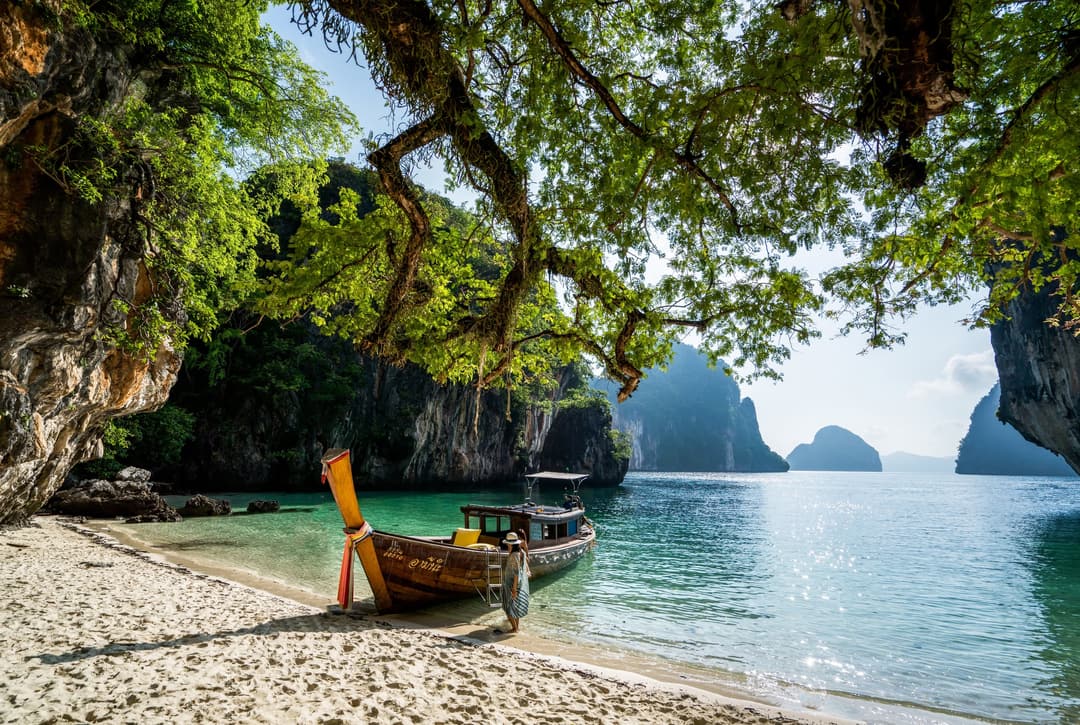 Wooden boat docked on a sandy beach, shaded by green trees, with clear blue water and limestone cliffs visible in the distance, Six Senses Yao Noi