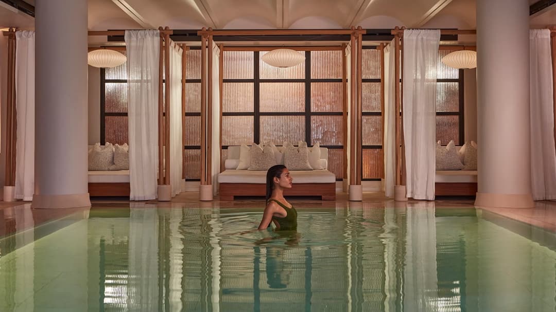 A woman wades in a calm indoor pool, with curtained lounge beds and soft warm lighting behind her, Claridge's