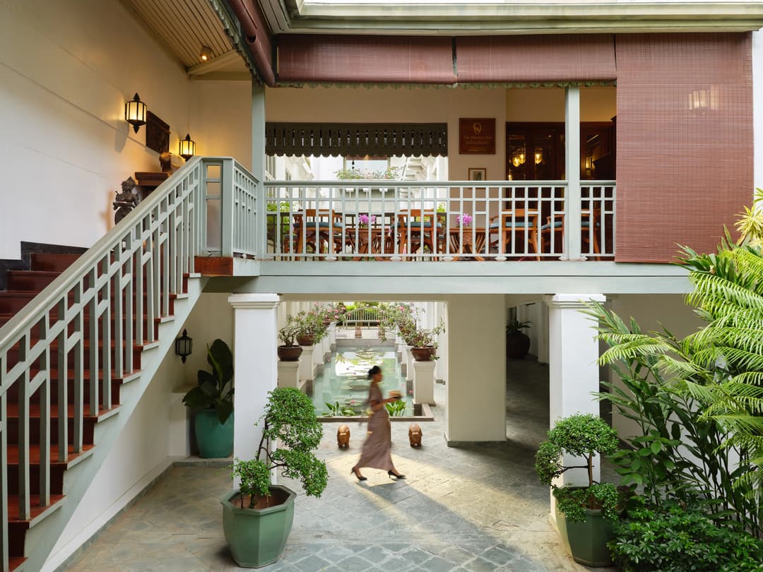 Colonial-style courtyard with white railings, lush plants, and a staff walking past a central reflecting pool, Mandarin Oriental Bangkok