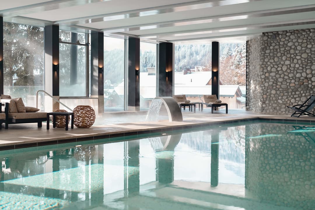 Modern indoor pool with curved water feature, stone wall, lounge chairs, and floor-to-ceiling windows overlooking snowy mountains, Carlton Hotel St. M