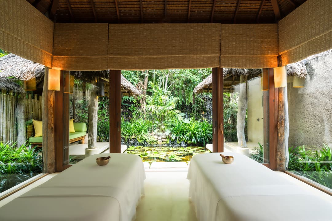 Open-air spa room with massage tables, garden and pond views, warm wooden details, gentle light, and a calm atmosphere inspired by nature, Six Senses