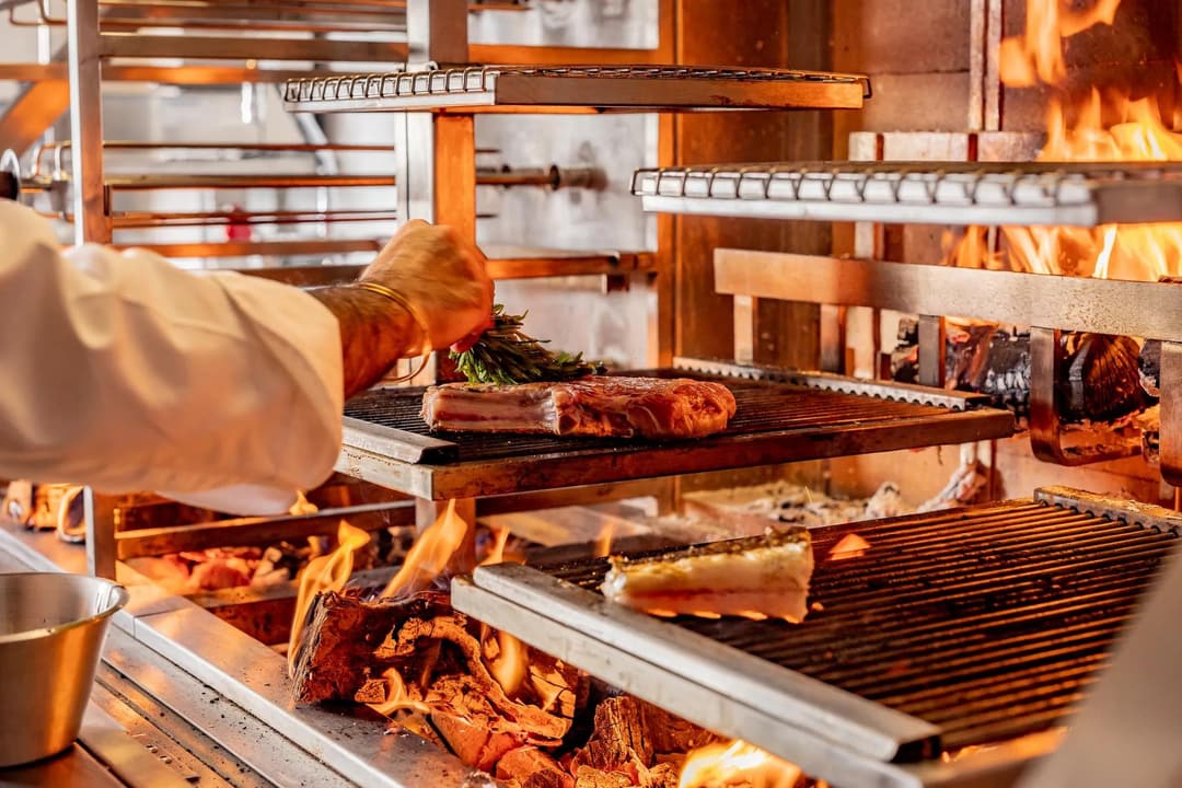 Chef placing herbs on meat cooking over open flames on a multi-level wood-fired grill, The Connaught