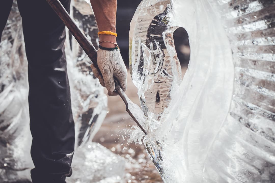 Person wearing work gloves using a chisel to carve or sculpt a large block of ice, with ice shavings visible, Carlton Hotel St. Moritz