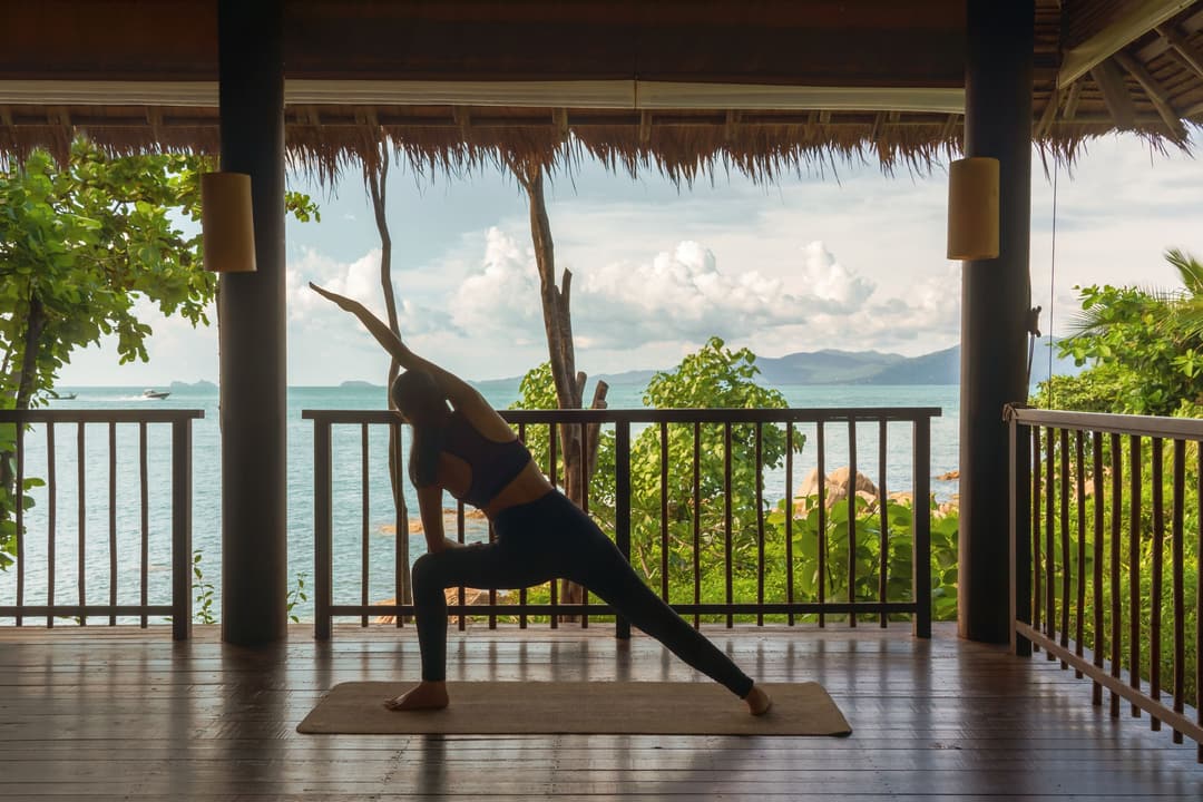 A guest enjoys a yoga lunge pose on a wooden deck with ocean views, shaded by a thatched roof and framed by greenery, Six Senses Samui