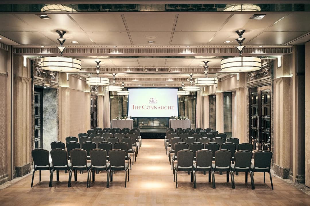 Conference room with rows of chairs facing a screen displaying The Connaught, under warm lighting and elegant detailing